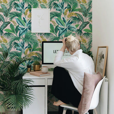 A college student sitting at their desk in dorm room in front of a wall featuring tropical peel and stick wallpaper. #color_pink-botanical