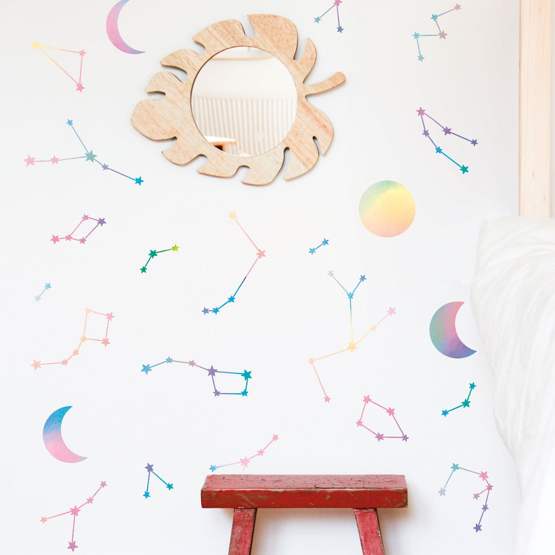 A red stool and a leaf-shaped mirrored next to a wall with holographic Constellations & Moons wall decals from Tempaper.