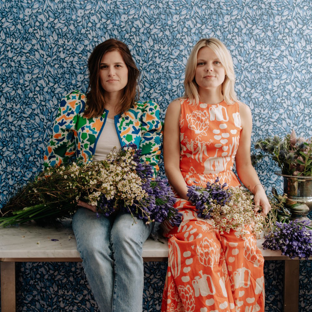 Tempaper's Flamboyan Peel And Stick Wallpaper By She She shown with two women holding plants.